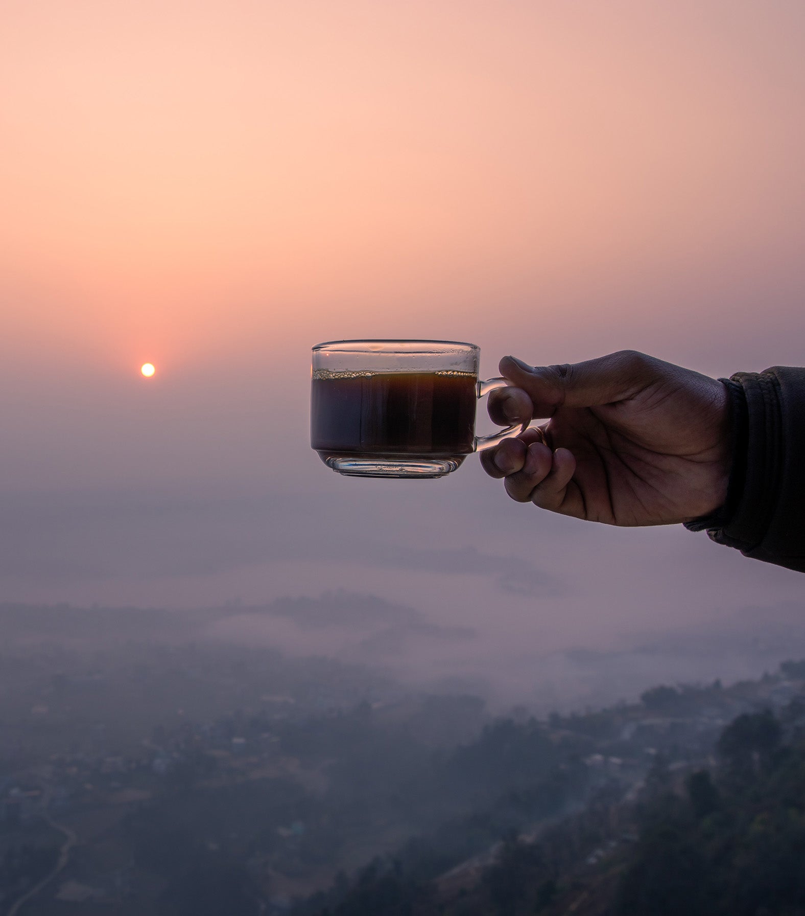 Hand holding a glass of coffee against a sunset sky with a cityscape below
